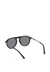 Lunettes de soleil à monture noire avec verres ronds foncés et branches métalliques fines, présentées en angle sur un fond blanc.
