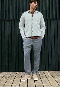 Hellgrauer, strukturierter Cardigan mit fünf Knöpfen, getragen über einem rostfarbenen Hemd, kombiniert mit grauen Hosen und beigen Sneakers, stehend auf einem Holzboden.