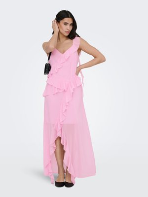 Femme en robe maxi rose sans manches à volants avec un bas asymétrique, sac à épaule noir et ballerines noires, posant devant un fond uni.