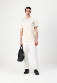 T-shirt en coton crème avec un petit graphique sur la poitrine gauche, associée à un pantalon blanc et un sac fourre-tout en cuir noir. Baskets noires.