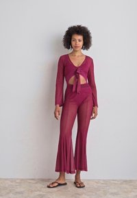 Conjunto de dos piezas burdeos que incluye un top corto de manga larga con lazo en la parte delantera y pantalones de tiro alto, acampanados y translúcidos. Sandalias negras planas.
