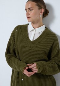 Olíva zöld, oversized kötött pulóver V kivágással, bordás textúrával és rojtos ujjak végével. Fehér galléros ing fölött viselve, amelyen látható egy lyuk.