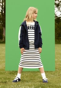 Bambino con giacca a track navy con accenti bianchi, vestito a righe bianco e navy, calze bianche e sneaker navy, in piedi sull'erba.