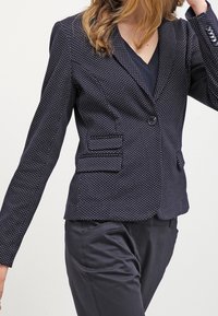 Marinblå blazer med vita prickar, enkel knappstängning, två framfickor och figursydd passform. Slät tygtextur.