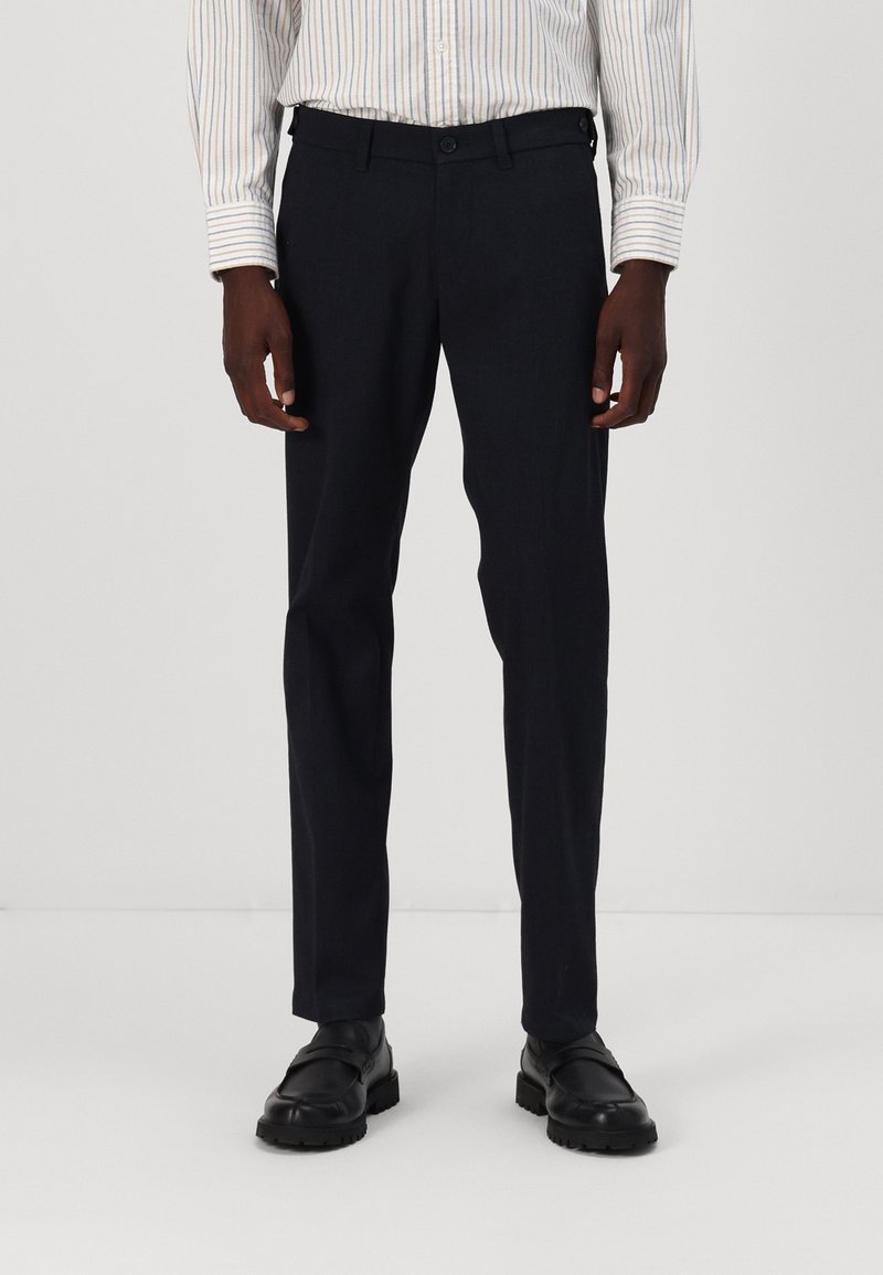 Pantalons sombres pour hommes avec une coupe ajustée, fabriqués dans un tissu lisse. Comprennent des poches avant et une fermeture à boutons, associés à des chaussures noires.