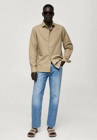 Beige skjorte med knaplukning, lyseblå lige bukser og brune sandaler. Modellen bærer mørke solbriller og fremviser et afslappet outfit.
