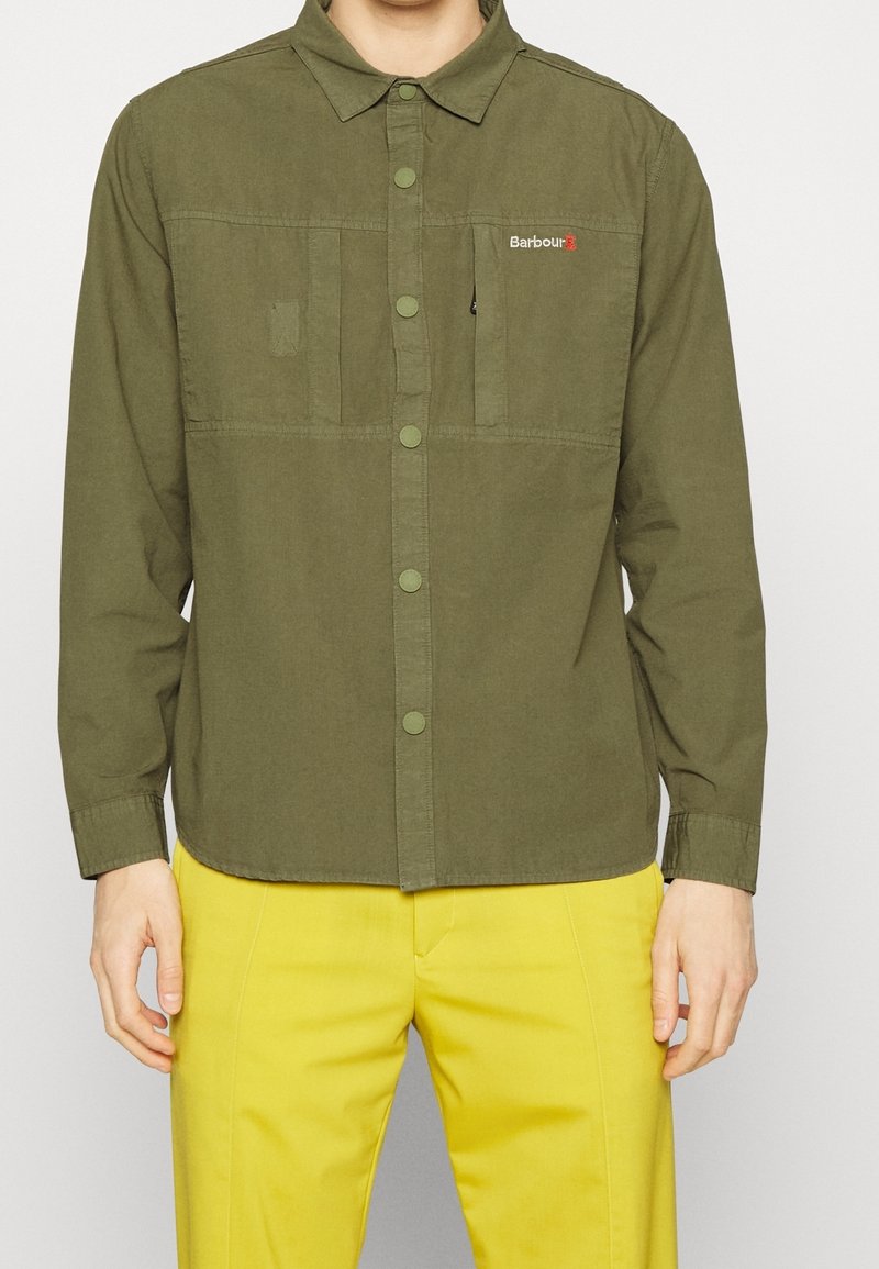 Homme portant une chemise boutonnée vert olive avec des poches poitrine et un pantalon jaune, se tenant devant un fond uni.