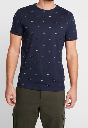 Mann trägt ein eng anliegendes marineblaues T-Shirt mit kleinen gelben Krabbenmotiven und olivgrünen Cargohosen, steht mit entspannten Armen.