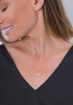 Goldkette mit einer dünnen Kette, die einen Ankh-Anhänger trägt, getragen auf einem nackten Hals, der sich von einem schwarzen V-Ausschnitt-Oberteil abhebt.