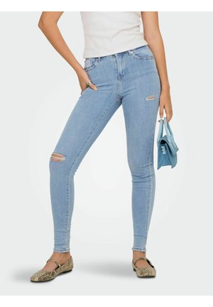 Persona che indossa jeans skinny azzurri strappati, ballerine beige con stampa serpente, top bianco senza maniche, che tiene una piccola borsa blu.