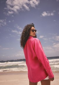 Pink linen shirt with long sleeves, featuring a collar and button details. Worn with shorts, set against a beach backdrop.