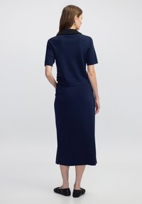 Žena s dlouhými vlasy, oblékající námořnickou modrou blůzu s krátkými rukávy a odpovídající midi sukni, otočená zády na jednobarevném světlém pozadí.