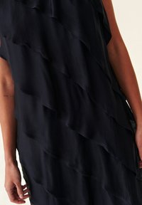 Robe noire sans manches avec des volants diagonaux superposés, portée par une personne avec un tatouage visible sur l'avant-bras gauche.