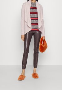 Hellrosa drapierte Strickjacke über einem grau gestreiften Pullover mit roten Akzenten, kombiniert mit braunen Kunstlederhosen und orangefarbenen Fellslippern. Orangefarbene Clutch.