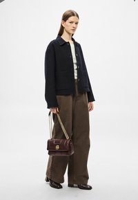 Casaco azul-marinho com botões e dois bolsos frontais, combinado com calças largas castanhas. Segura uma mala de mão em pele bordô com alça em corrente dourada.