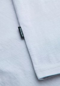 Vaaleansininen t-paita, pehmeää puuvillamateriaalia, lyhyet hihat, joissa musta "ombre" -logotarra sauman kohdalla. Sujuva pinta, minimaalinen muotoilu.