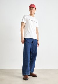 T-shirt blanc à manches courtes avec le logo noir "TOMMY HILFIGER", associé à un jean en denim large bleu et des chaussures slip-on marron. Casquette rose sur la tête.