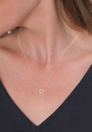 Guldkæde med en delikat kæde, der har et hjerteformet vedhæng prydet med små klare krystaller, sat mod en mørk baggrund.