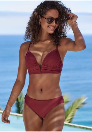 Žena s kučeravými vlasmi, ktorá nosí slnečné okuliare a tmavočervené bikiny, sa usmieva vonku pri bazéne a oceáne pod jasnou oblohou.