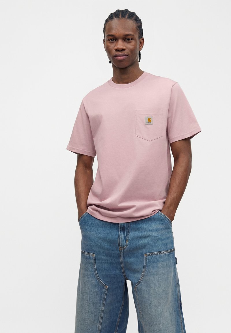 Young man with braided hair wearing a light pink short-sleeve t-shirt with chest pocket and loose blue denim jeans, standing with hands in pockets.
