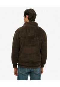 Brun fleecejakke med høj krave, tekstureret stof og ribbede manchetter; har en rygklap og en afslappet pasform. Denimjeans bæres.