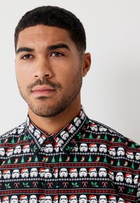 Lyhythihainen napillinen paita, jossa on musta pohja ja toistuva kuvio, jossa on Sokertajia, joulukuusia, karamellikeppejä ja puna-vihreää piikkipensasta.