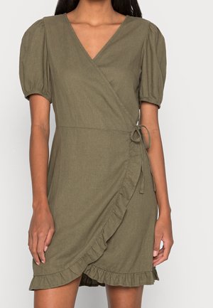 Femme portant une robe portefeuille vert olive avec manches bouffantes, bordure à volants et fermeture à nouer sur le côté, vue des épaules jusqu'à mi-cuisse.