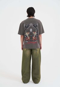 Grå grafisk t-shirt med et detaljeret print af slanger, fugle og et kors på ryggen; parret med oversized grønne denimbukser.