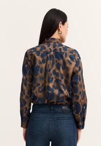 Woman with straight dark hair wears blue and brown leopard print long-sleeve shirt tucked into dark blue jeans, facing away.