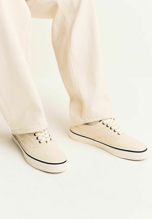 Asmuo, dėvintis smėlio spalvos drobinius sportbačius su juodais apvadais ir raišteliais, derinamus su šviesiai smėlio spalvos kelnėmis, ant balto fono.