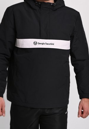 Muž v černé bundě Sergio Tacchini s bílým horizontálním pruhem a logem přes hrudník, stojící s uvolněnýma rukama.