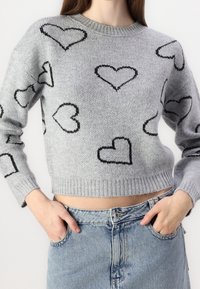 Světle šedý pletený krátký svetr s černě obrysy srdíčkových vzorů, nošený s světle modrými high-waisted džíny, s rukama v kapsách.