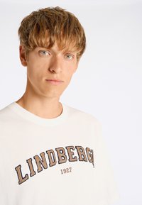 Camiseta de algodón blanca con mangas cortas, que presenta "LINDBERG 1927" en letras bordadas en azul marino y beige en el pecho.