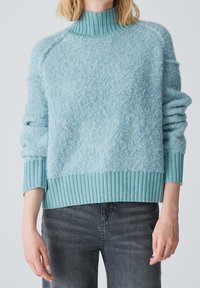 Femme portant un pull tricoté bleu clair à texture avec un col roulé, des poignets et un ourlet côtelés, associé à un jean gris foncé.