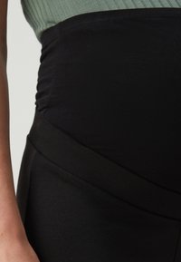 Vêtement noir ajusté avec des détails froncés sur les côtés, présentant une texture lisse et une large ceinture créant une silhouette structurée.