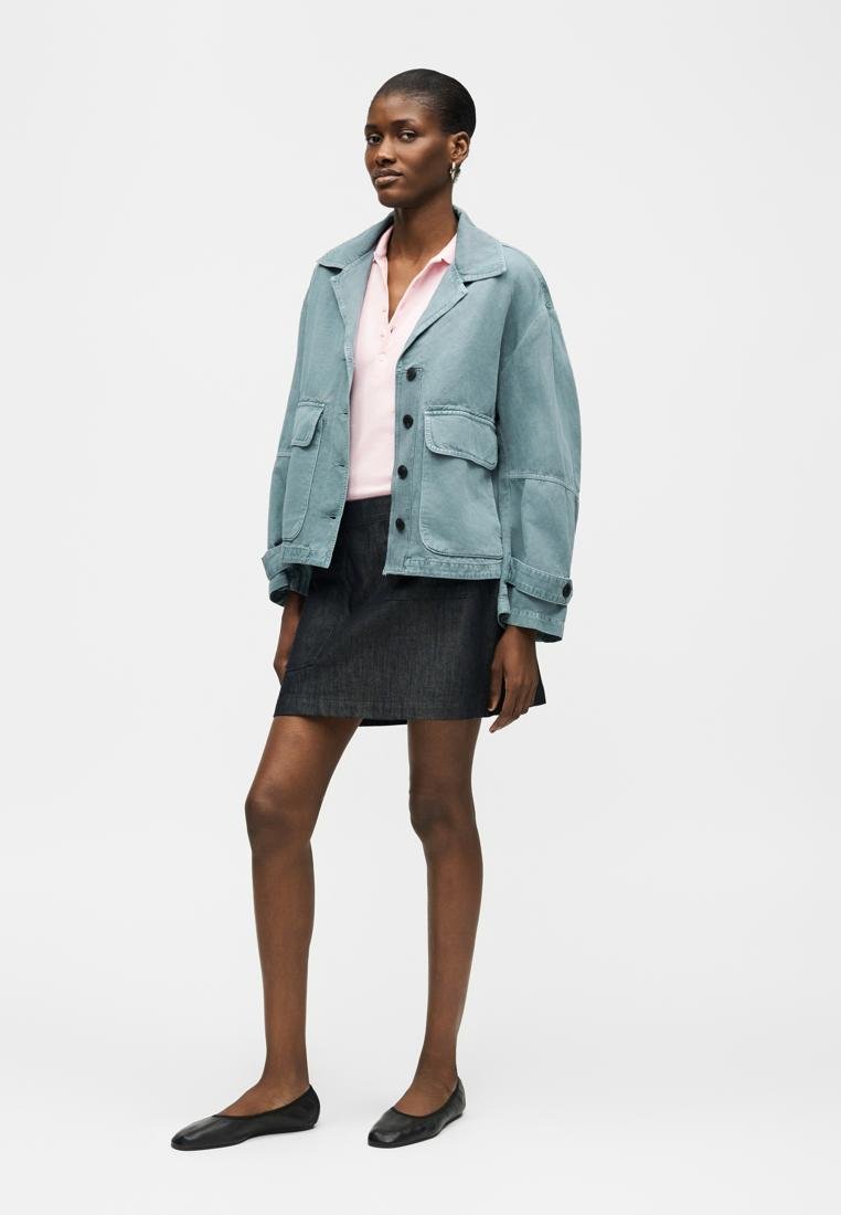Femme portant une veste bleu clair surdimensionnée, une chemise rose pâle, une jupe gris foncé et des ballerines noires, debout devant un fond blanc.