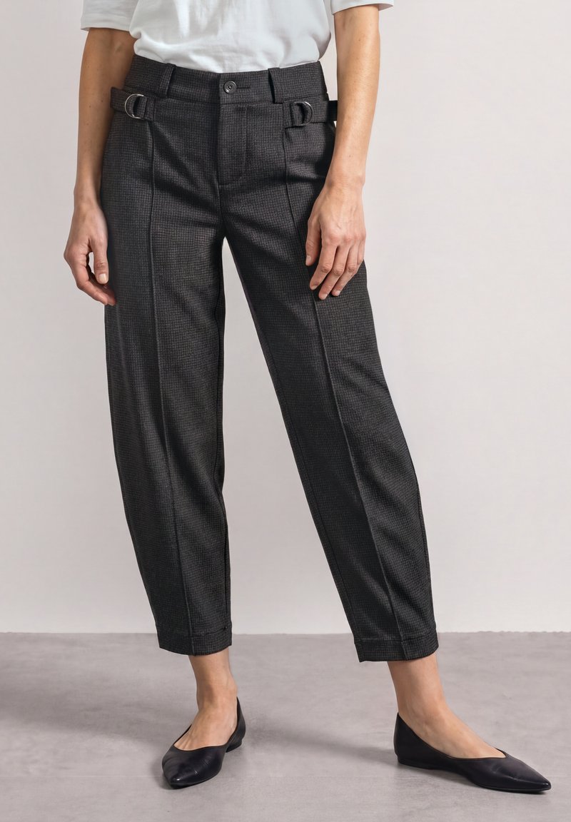Femme portant un pantalon court gris foncé taillé sur mesure avec des détails de boucle latérale et des ballerines noires pointues, debout sur un sol gris.