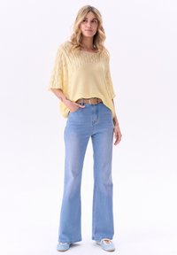 Lichtgele gebreide top met een losse pasvorm en een open patroon, gecombineerd met lichtblauwe flared jeans en blauwe flats, aangevuld met een bruine riem.