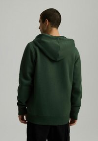 Ciemnozielona bluza z kapturem, wykonana z miękkiego, teksturowanego materiału. Posiada pojemny kaptur i ściągacze, z prostym, minimalistycznym designem.