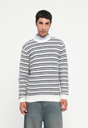 Pulover tricotat alb și negru cu un model zigzag, guler rotund și mansete cu borduri. Asortat cu o cămașă cu guler de un bleu deschis și blugi.