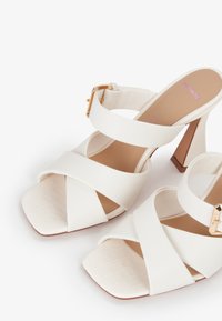 Witte high-heeled sandalen met een vierkante neus, voorzien van kruislings gespen en een enkelbandje met gouden gesp. Textuur van de voetbed met een subtiel krokodillenpatroon.