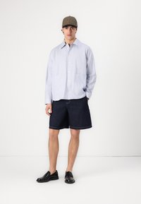 Lys blå og hvit stripete skjorte med knapper, mørk denimshorts og svarte slip-on sko; har en rutete caps og to frontlommer.