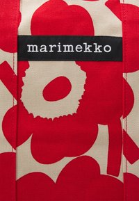 Sac fourre-tout Marimekko avec un motif floral rouge sur fond beige, en toile résistante, et une étiquette logo noire.
