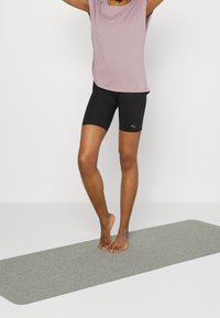 Camisa deportiva de color púrpura claro y pantalones cortos negros en un modelo que está de pie sobre una esterilla de yoga gris, mostrando la textura del tejido y un diseño simple.