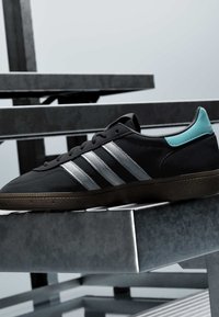 Baskets Adidas noires avec des bandes argentées et une languette bleu clair au talon reposant sur un banc en métal gris avec un arrière-plan flou.
