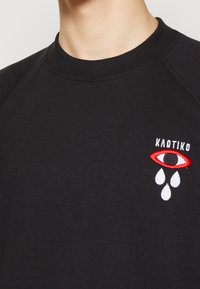 Svart sweatshirt med ribbad rundhals, prydd med broderad öga och droppdesign i röd och vit på vänster bröst. Slät textur.