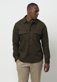 Camicia a maniche lunghe colore verde oliva scuro, realizzata in tessuto testurizzato, con due tasche sul petto e un colletto.