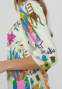 Bluză florală multicoloră, realizată dintr-un material moale, cremos. Designul include flori vibrante, pensule și textul "ART studio" în negru.
