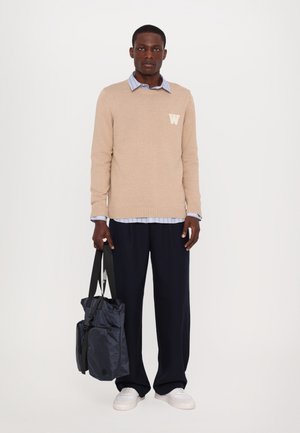 Beige genser med et "W"-logo, lagd over en lyseblå skjorte med krage. Mørk marineblå vid bukser og en svart og grå tote bag. Hvite joggesko.