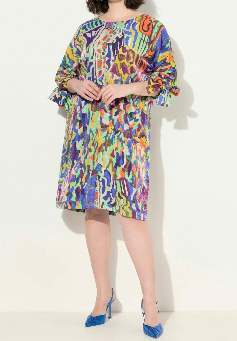 Femme portant une robe mi-longue à motifs abstraits multicolores avec des manches trois-quarts et des chaussures à talons bleues à bride arrière.